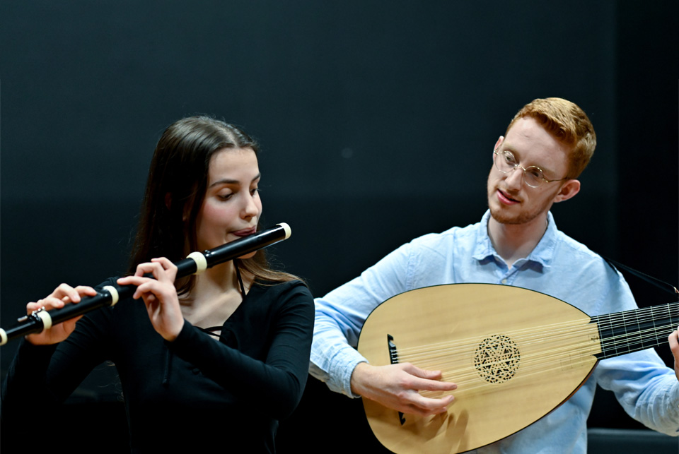 A male student, wearing a blue shirt, playing the lute, with a female student, wearing a dark shirt, performing the historical flute.